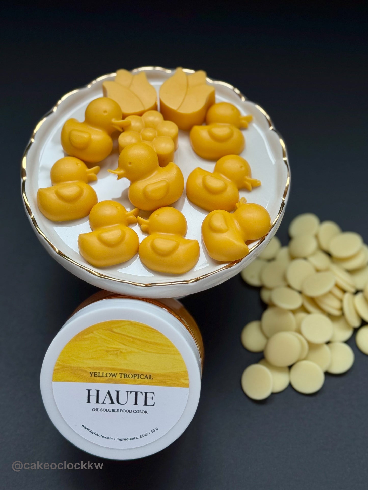 Yellow duck-shaped chocolates in a container with 'Haute' food color packaging on a dark background and white chocolate callets. Best Haute Food Colors by Ksenia Penkina, Natural Colouring, Water Soluble, Oil Soluble, Fat Soluble Powder, Professional, Chefrubber, Colourmill, Chefmaster, Silikomart, Pavoni, Decorelief, Matfer, PastryClass, Valrhona, Cacao Barry, For Macarons, For Cakes